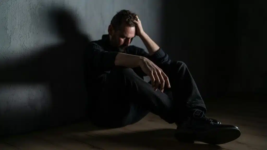 A man sits alone in a dark room with his hand on his head, looking stressed and sad.