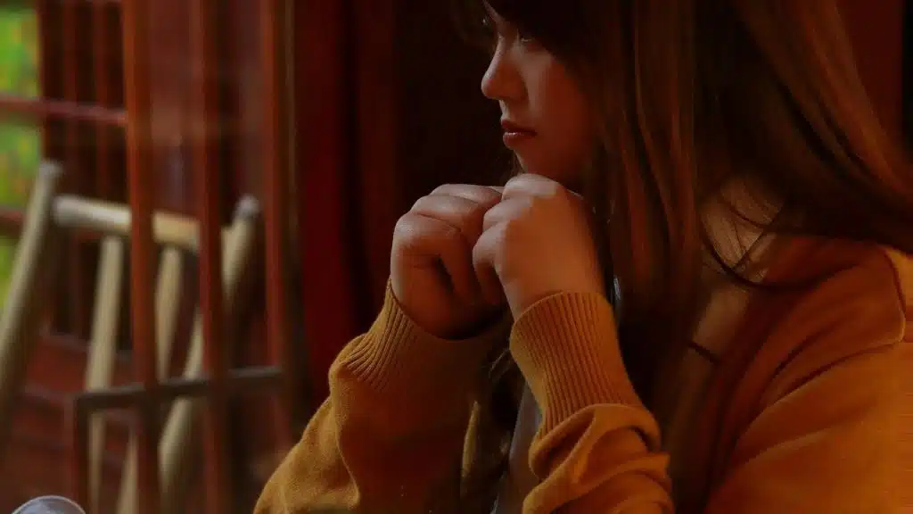 A person sits indoors with hands clasped near their chin, looking thoughtful.