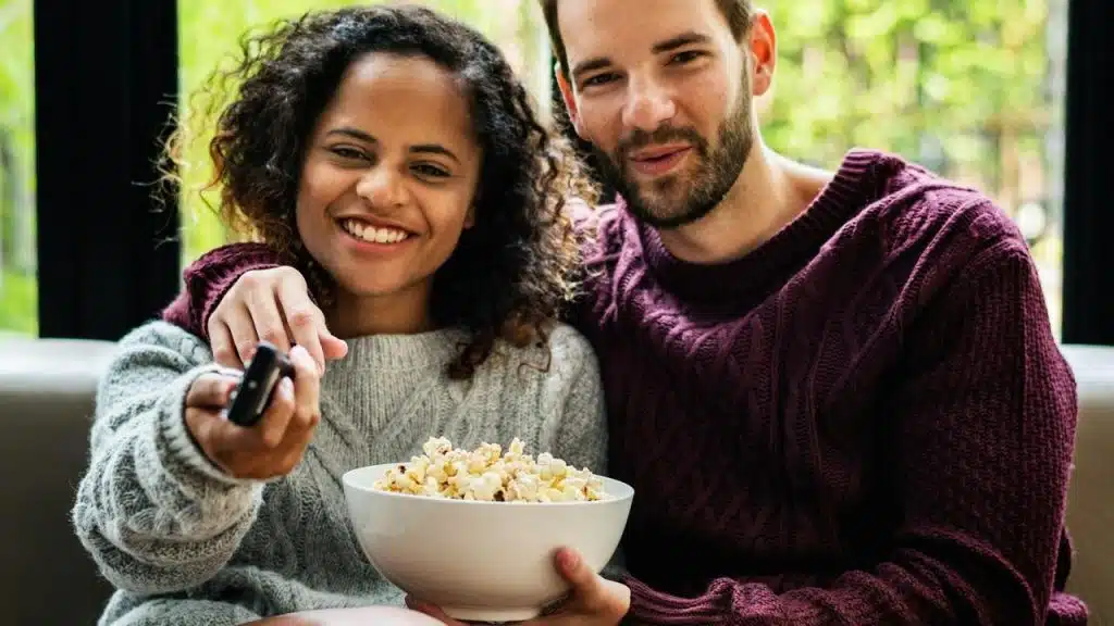 A couple smiling while watching TV and sharing popcorn.