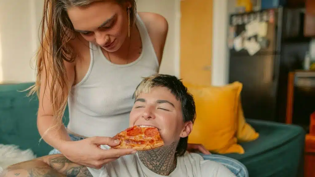 A woman sitting on a couch feeding a slice of pizza to a man who sits while leaning against her legs.