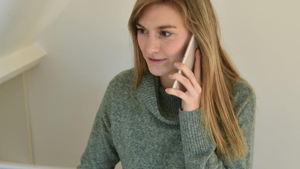 An indoor scene with a woman in a green sweater, holding a phone and speaking into it.