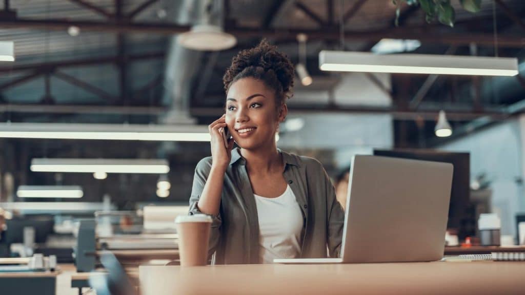 A woman sitting at a desk in an office, smiling and talking on a cell phone.