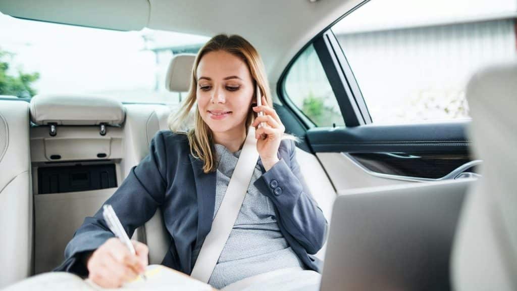 A man writing in a notebook while talking on the phone in the back seat of a car.