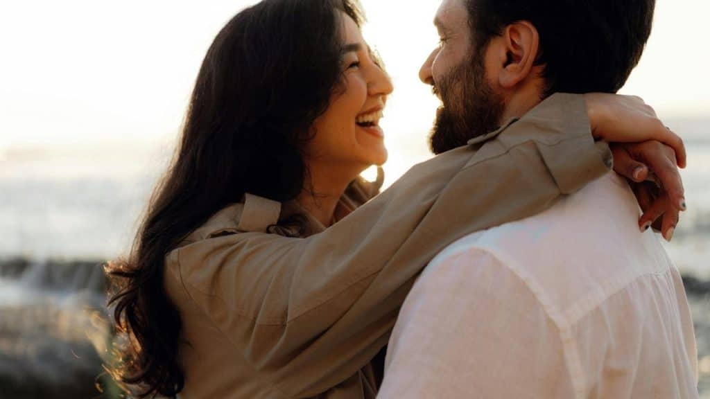 A happy couple standing in sunlight, holding hands and smiling at each other.