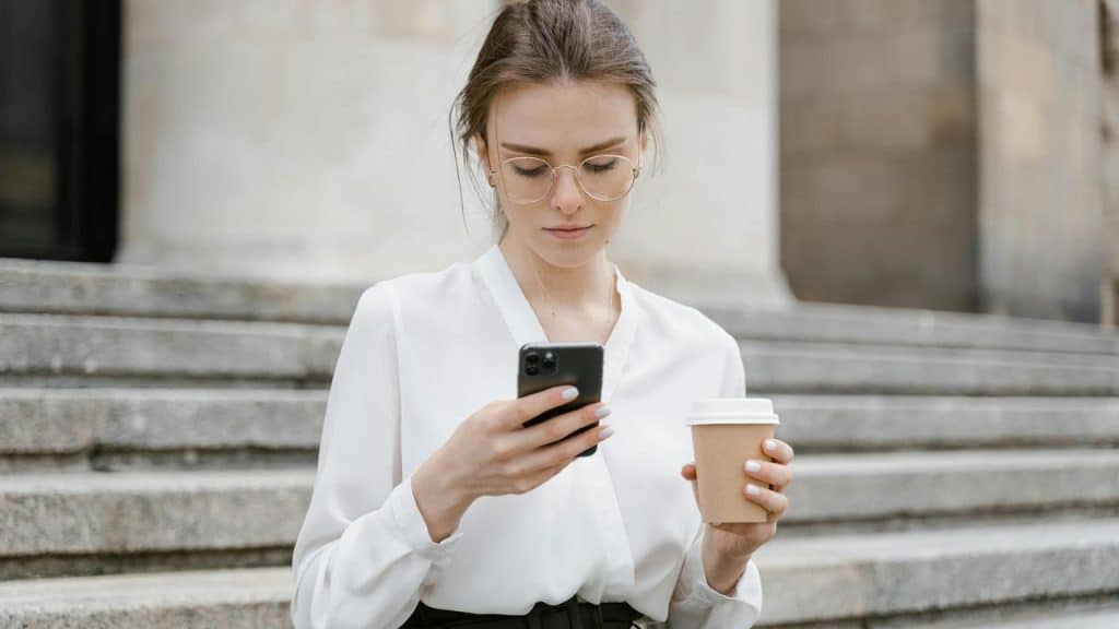 A woman with glasses looking at her phone while holding a coffee cup on outdoor steps.