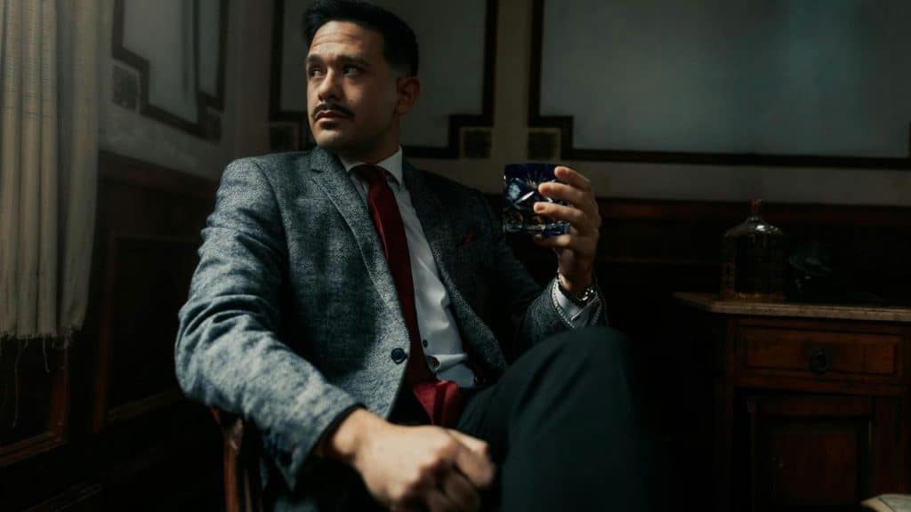 A man wearing a suit and holding a drink, standing with his arms folded and looking away.