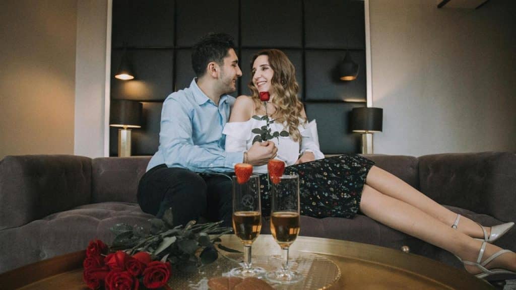 A man and woman sitting on a couch with roses and champagne in front of them.