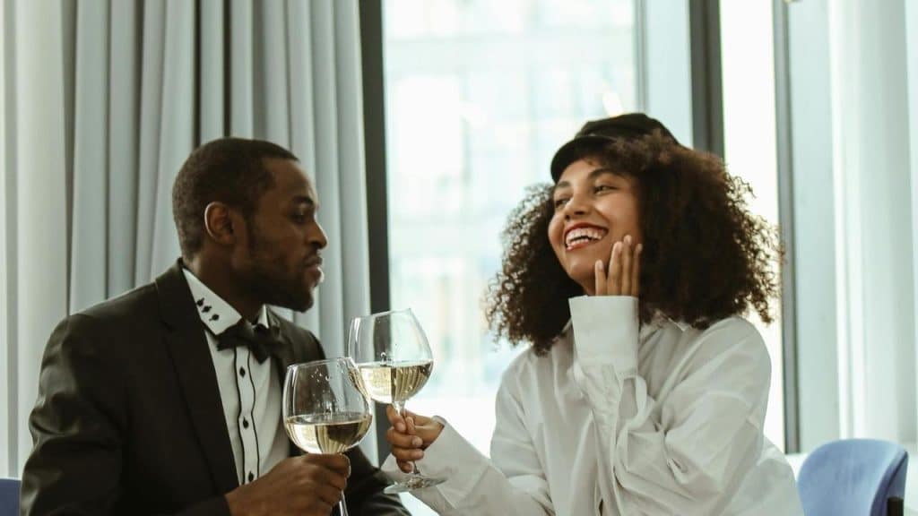 A man and woman dressed up in fancy attire, holding glasses of white wine and laughing.