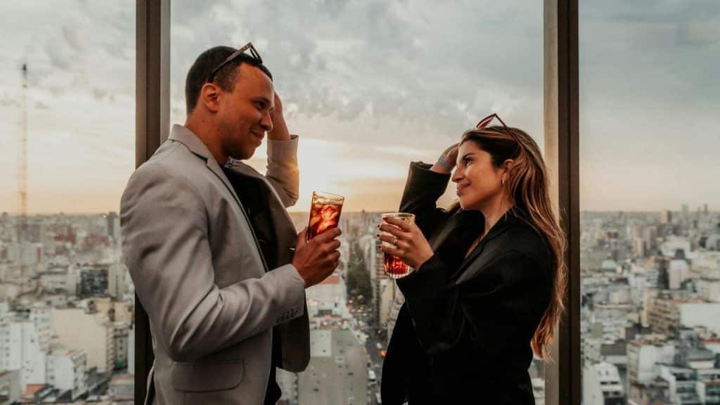 A man and woman standing on a rooftop drinking wine at dusk.