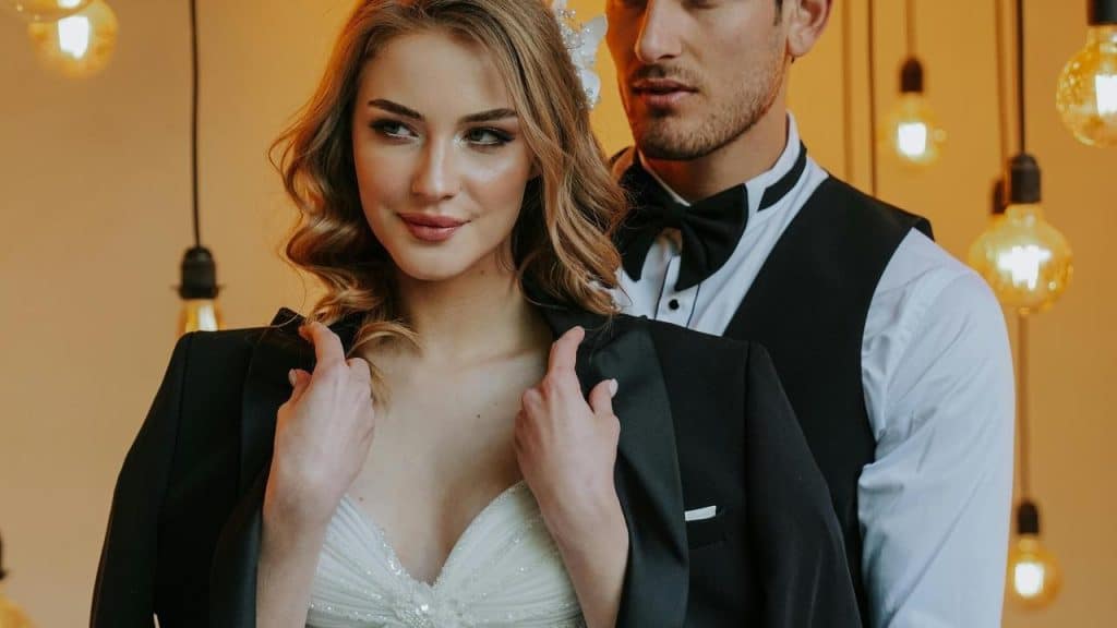 A couple dressed in formal attire standing together under warm hanging lights.