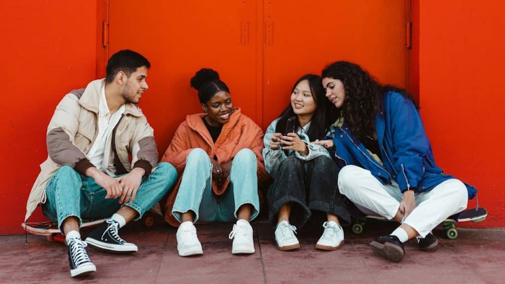 A group of friends sitting on the ground while leaning against a red wall and looking at a mobile screen that one of them is holding up.