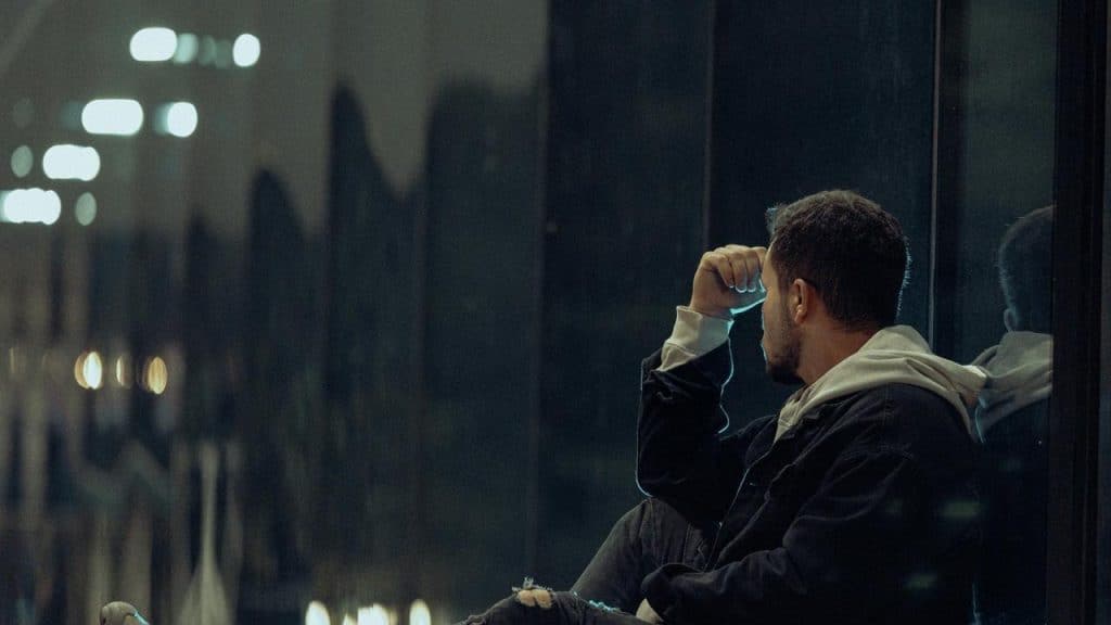 A man sitting on the ground, looking at a glass wall, his face contemplative, at night time.