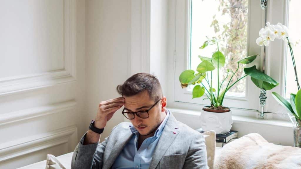 A man, seated on the couch with his hand on his forehead, appears to be stressed.