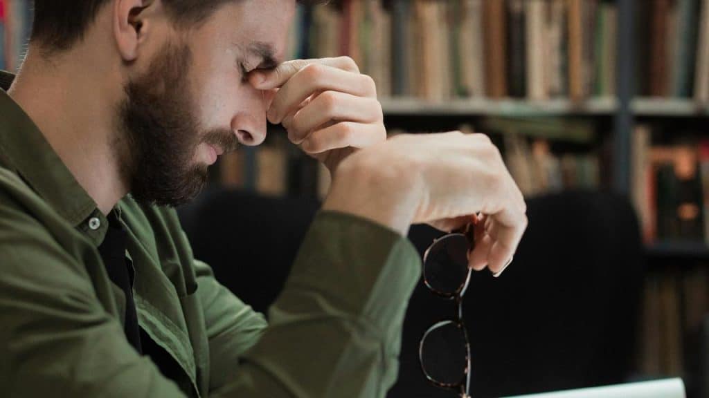 A man with his hands on his eyes and holding glasses at a desk.