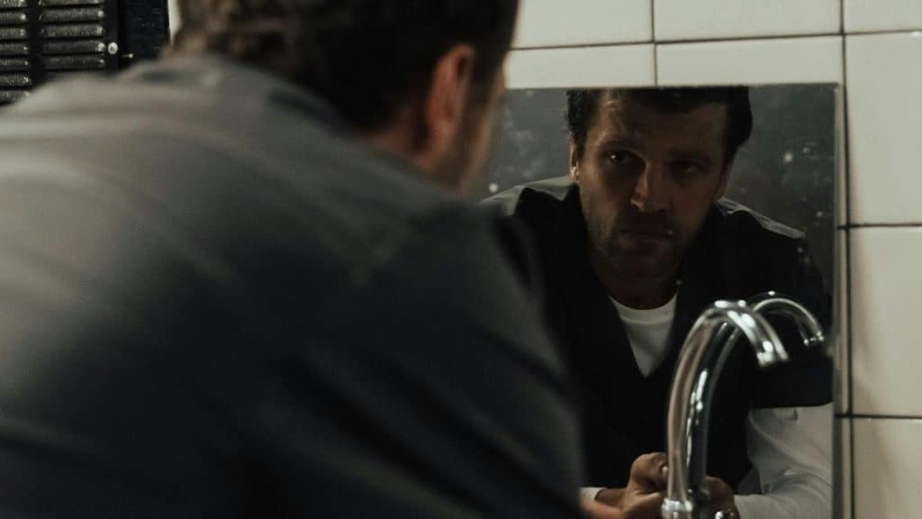 A man staring at his reflection in a dimly lit bathroom mirror.