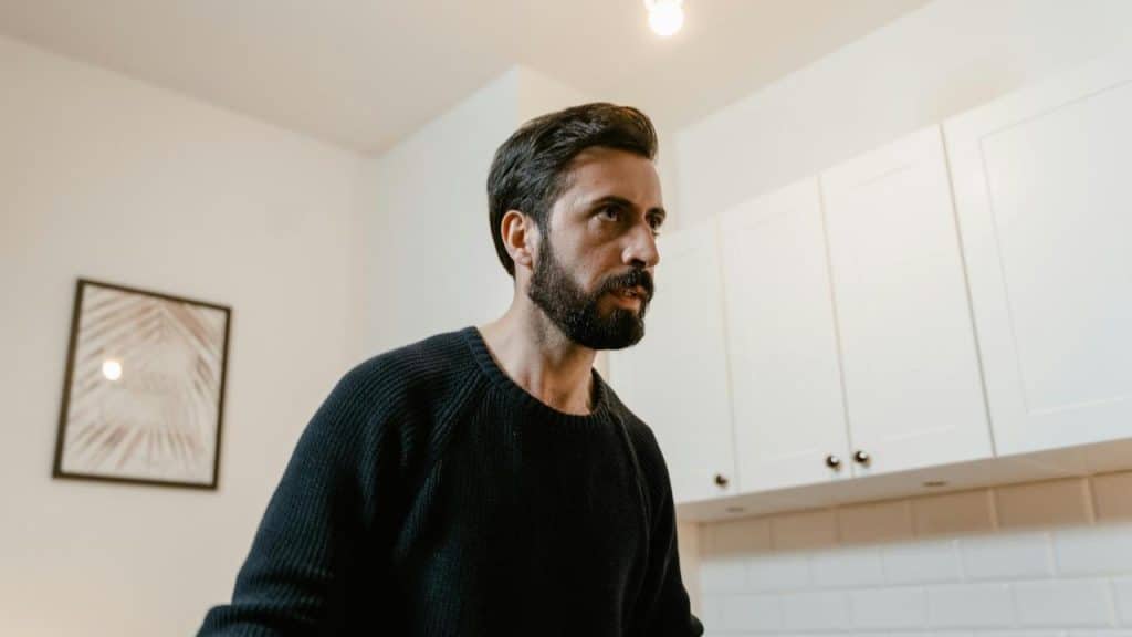 A bearded man standing indoors, looking to the side.