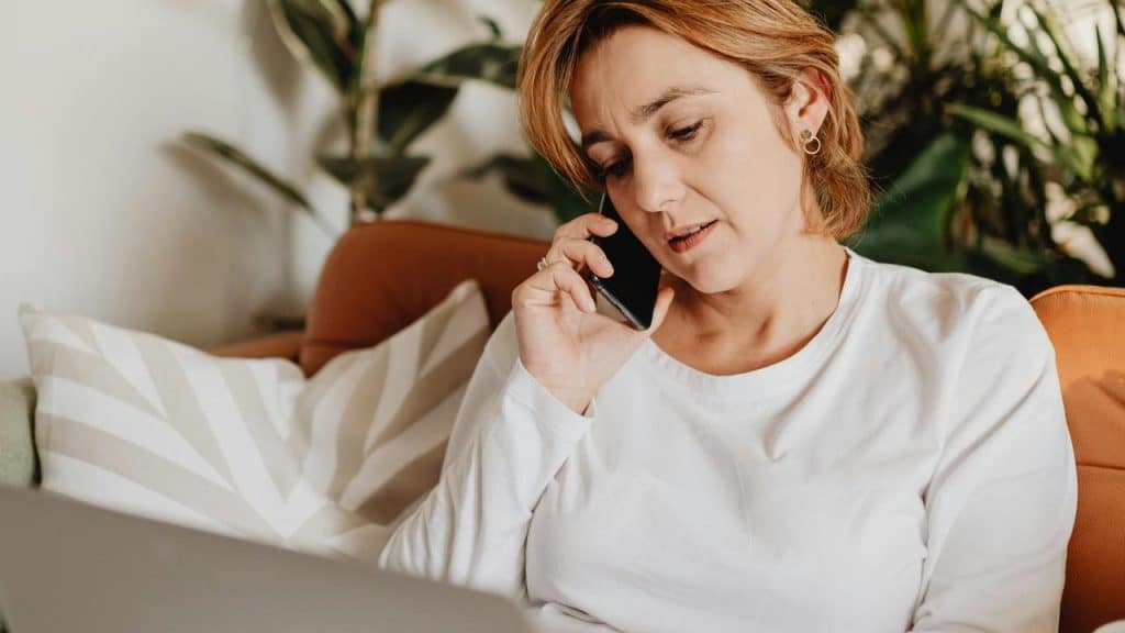 A woman talking on a phone while looking at a laptop on a couch.