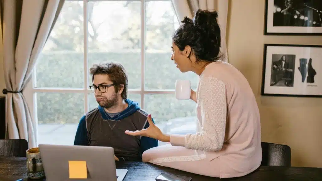 A woman gesturing while talking as a man sits at a laptop looking frustrated.