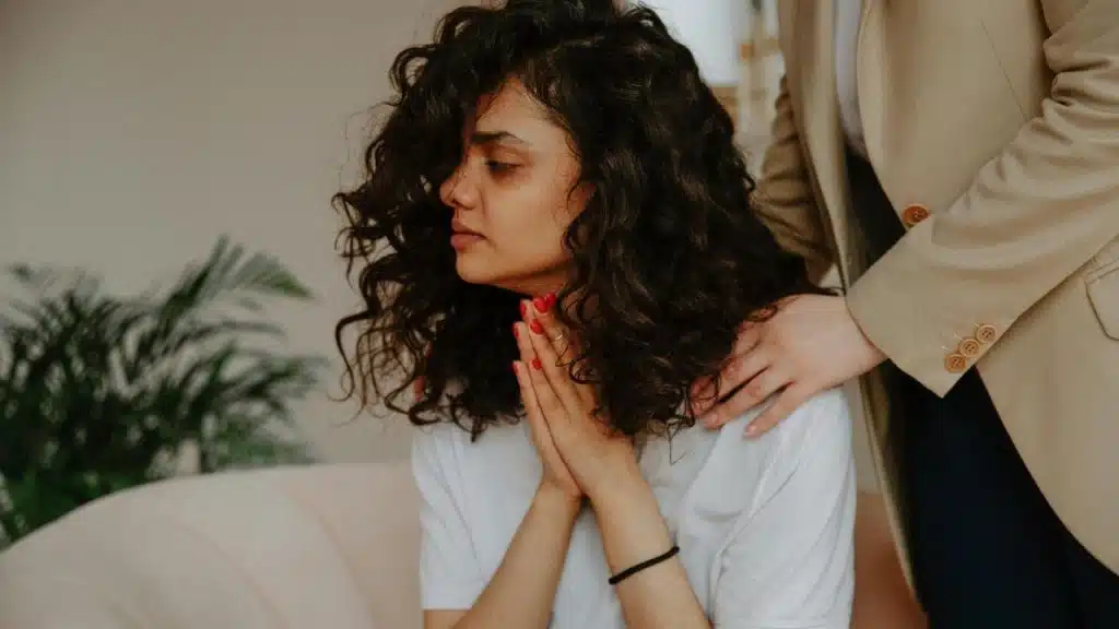 A woman with curly hair clasping her hands while another person touches her shoulder.