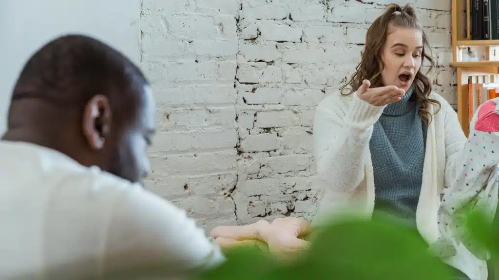 A woman reacting with surprise while holding up a piece of clothing as a man sits nearby.