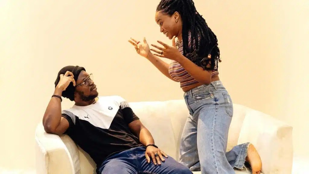 A woman gesturing animatedly while a man sits on a couch listening.