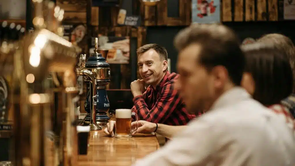 A man smiling at a bar with a beer while others sit nearby.