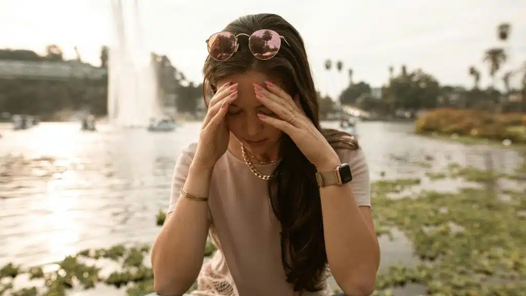 A woman holding her temples while sitting near a lake.