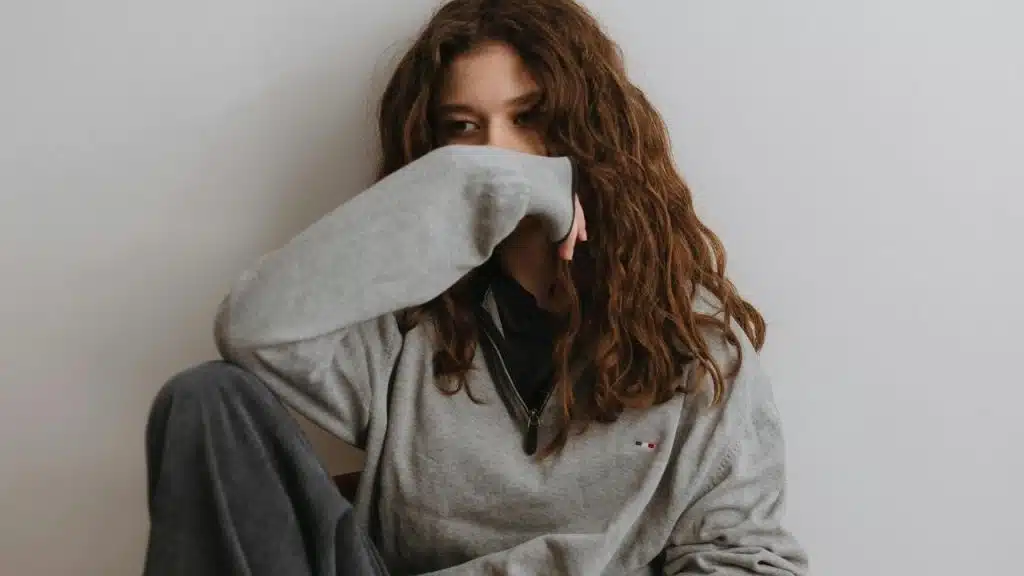 A woman covering her mouth with her sleeve while sitting against a wall.