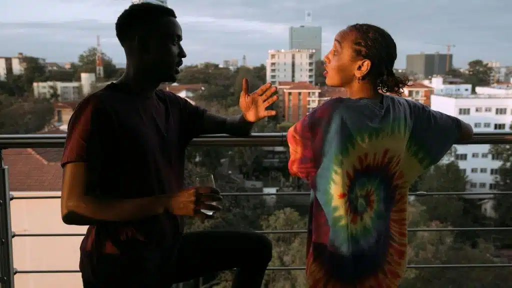 A man gesturing while talking as a woman reacts on a balcony.