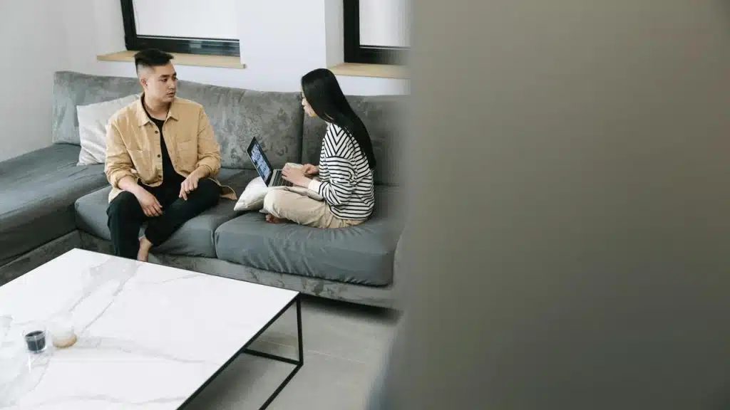 A couple sitting on a couch, talking while using a laptop.