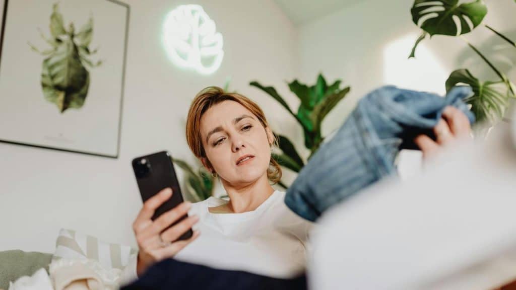 A woman holding a smartphone while examining a piece of clothing.