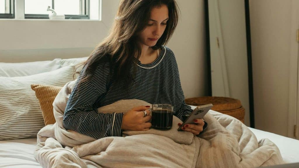 A woman in a striped shirt sitting in bed with coffee and her phone on a sunny morning.