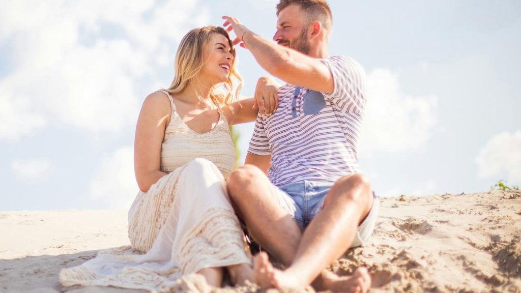A smiling couple sitting together on the sand enjoying a sunny day.