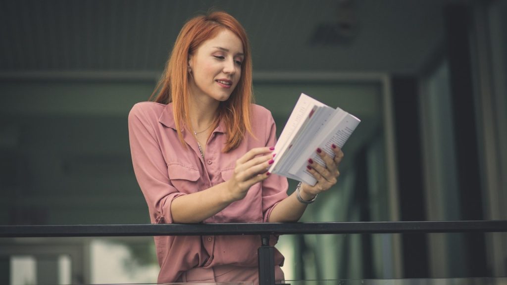 A woman reading a book while standing on a balcony.
