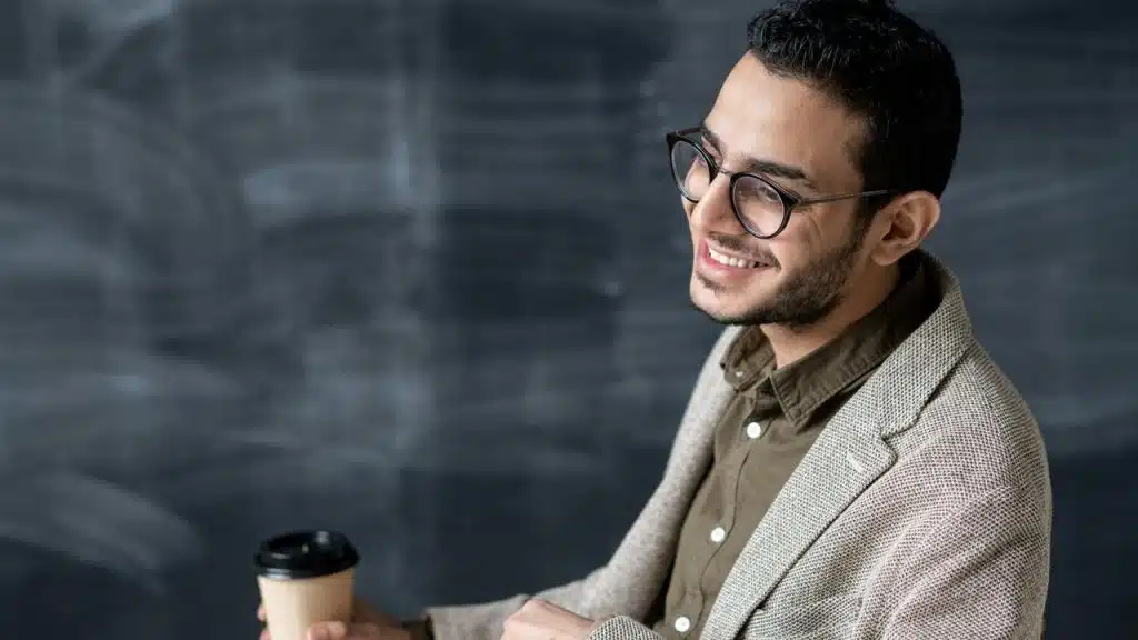 A man wearing glasses and a blazer smiling while holding a coffee cup.