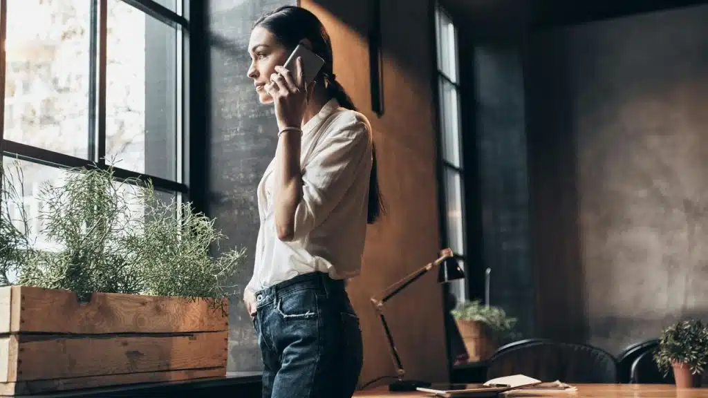 A woman standing by a window talking on the phone.
