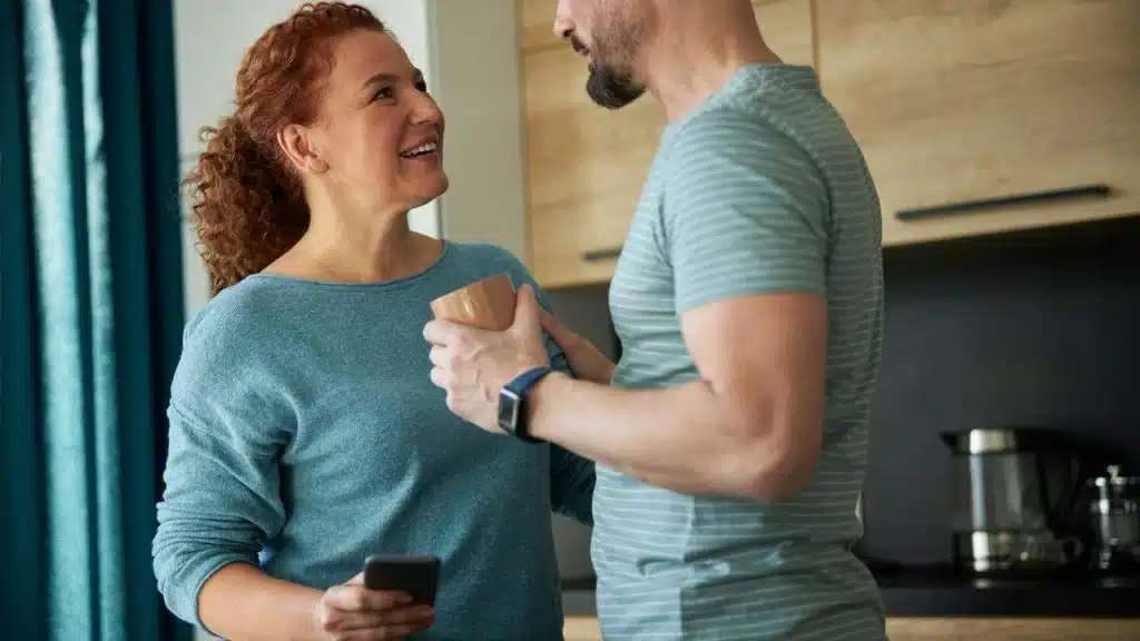 A smiling couple standing in a kitchen, holding a cup and a phone while talking.