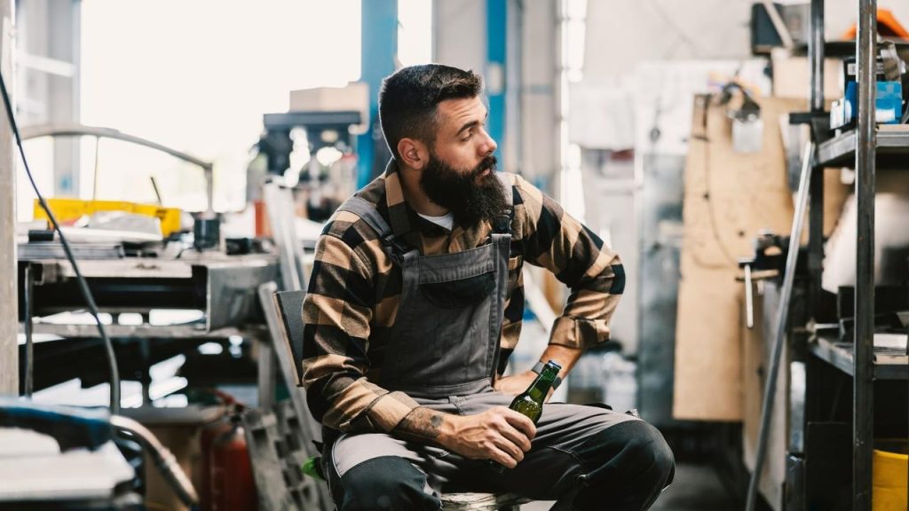 A man in work overalls sitting with a bottle in his hand.