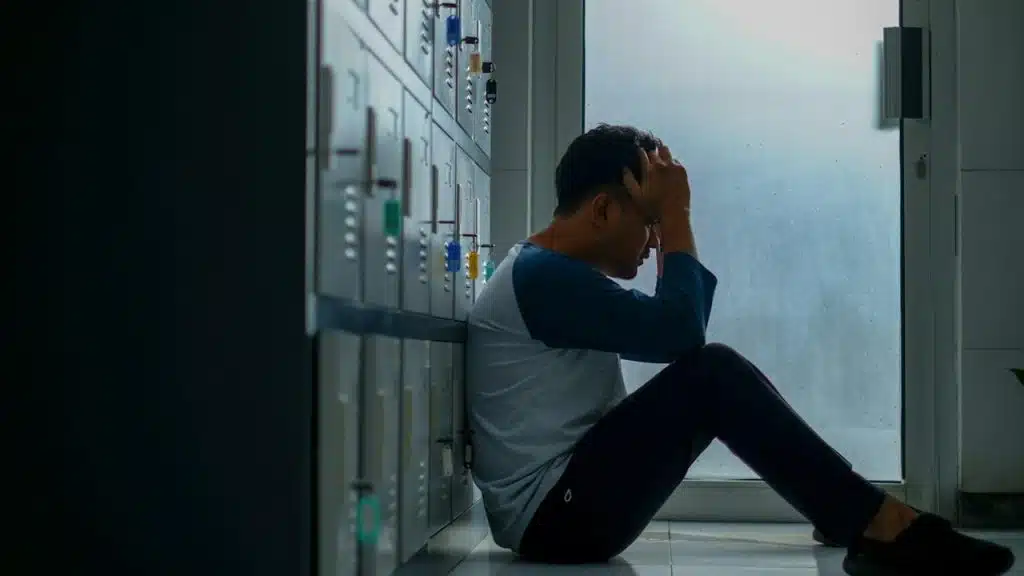 A man is sitting on the floor while leaning on some lockers while holding his head in his hands.