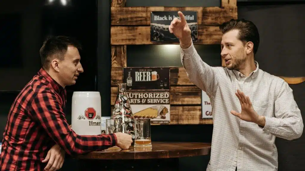 A man gesturing animatedly while talking with another person at a bar table.