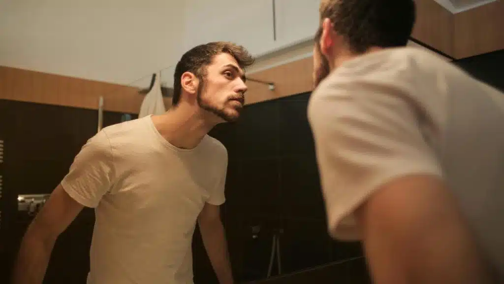 A man leaning toward a bathroom mirror, closely examining his face.