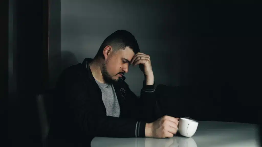 A man sitting at a table holding his head, looking stressed while holding a cup.