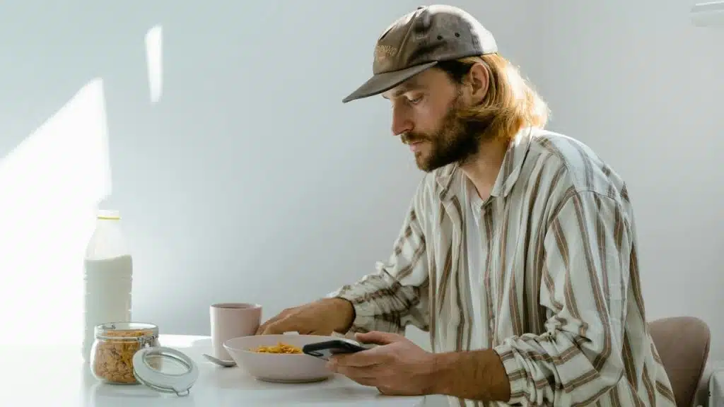 A man eating cereal at a table while looking at his phone.