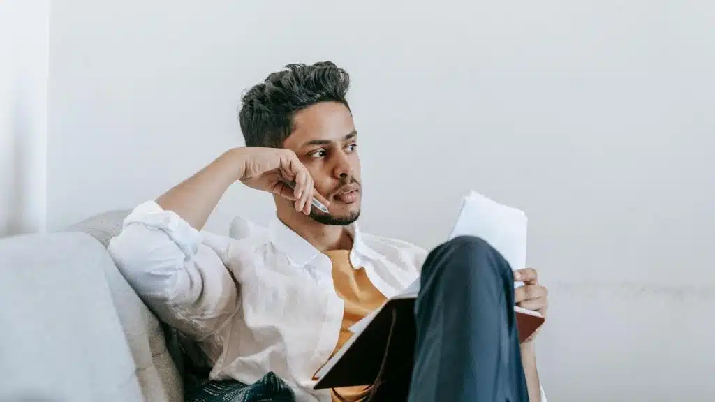 A man sitting on a couch holding a notebook, looking thoughtful.
