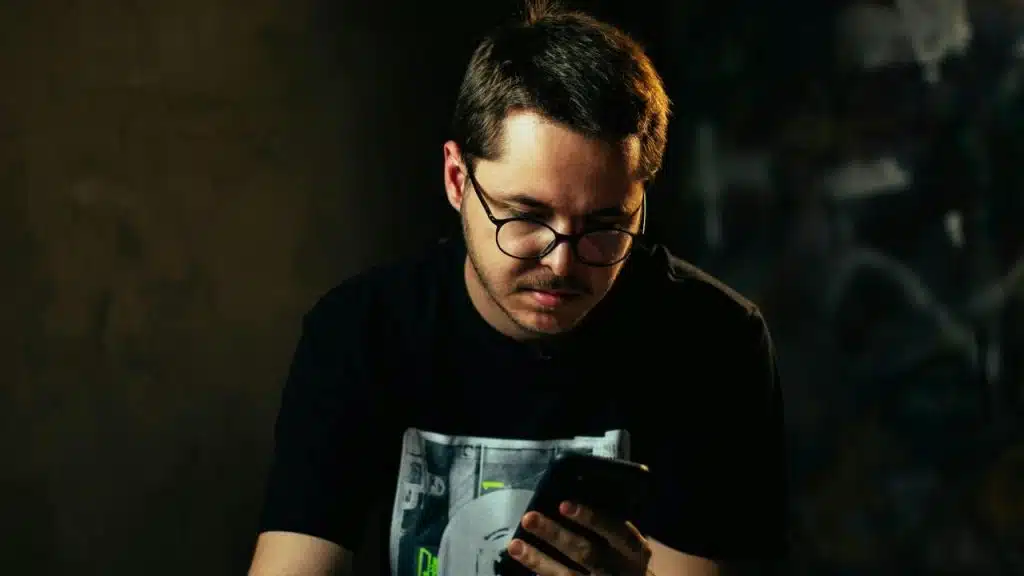 A man wearing glasses looking down at his phone in a dimly lit setting.