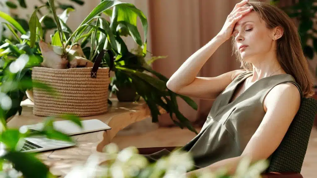 A woman sitting indoors near plants, resting her hand on her forehead.