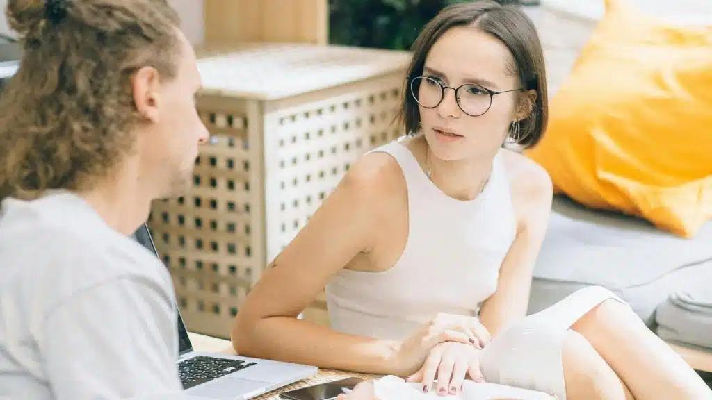 A woman wearing glasses talking with a man while sitting beside a laptop.