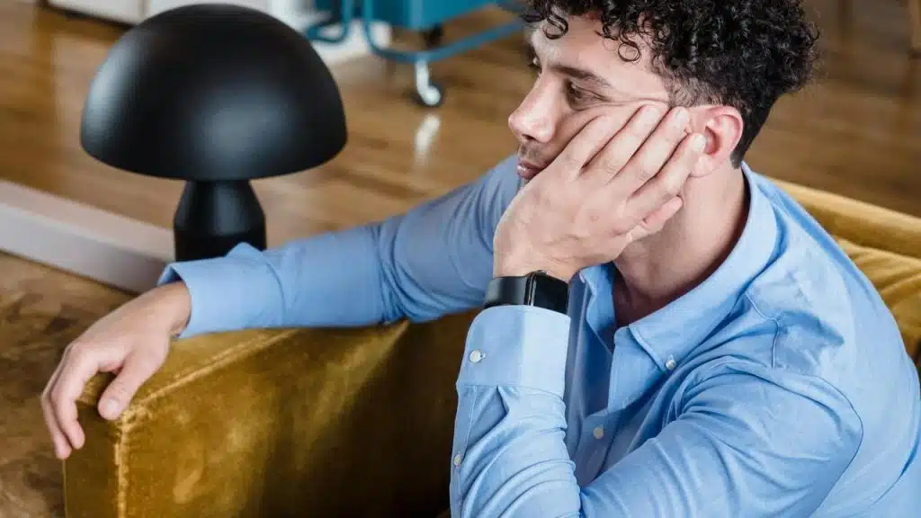 A man resting his head on his hand while sitting on a couch next to a lamp.