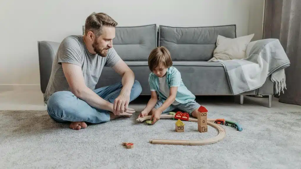 A man and a child sitting on the floor playing with a wooden toy train set.