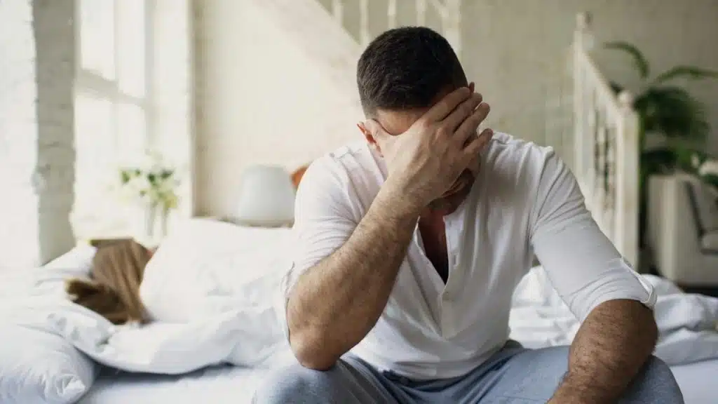 A stressed man is sitting on a bed while another person is lying in the background under the covers
